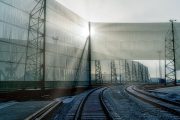 Railway tracks curve gently beside tall, translucent wind fences, illuminated by sunlight