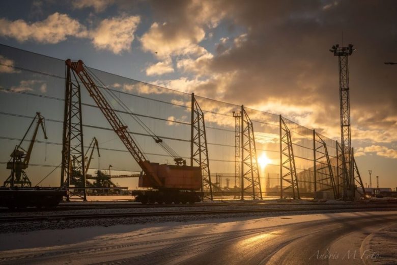A large crane sits on tracks behind a tall industrial wind fence at sunset