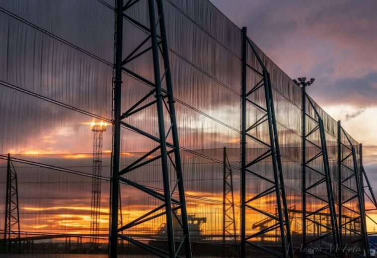 A large industrial fence structure stands against a vibrant sunset sky