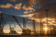 Cranes and industrial equipment silhouetted against a sunset behind a tall fence