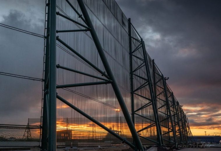 A large industrial fence structure with a sunset visible through its mesh panels