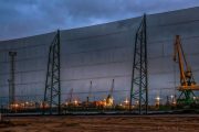 A large industrial wind fence stands in front of a port with cranes and ships