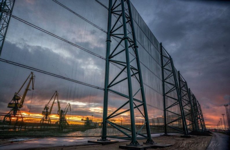 A large industrial wind fence stands against a dramatic sunset with cranes in the background
