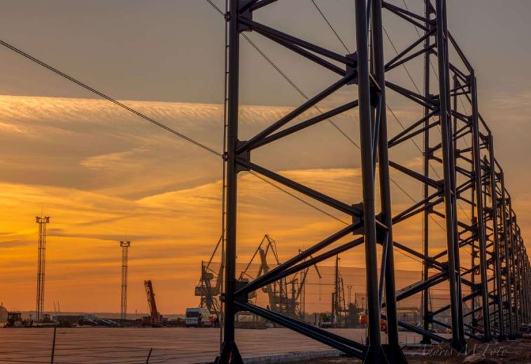 A metal structure stands tall against a vibrant sunset sky with cranes in the background
