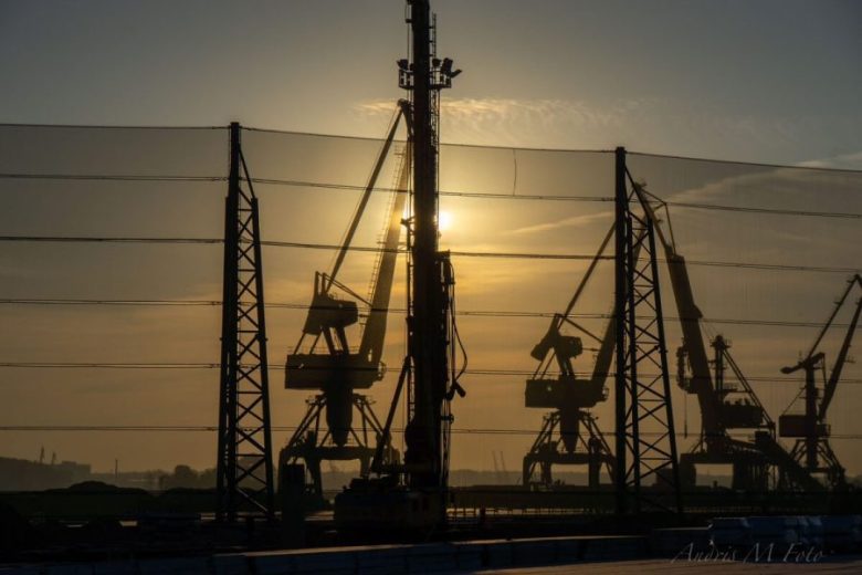 Silhouetted cranes and towers stand against a sunset, with a mesh fence in the foreground