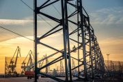 Steel framework structure with workers on lifts against a sunset sky at a port