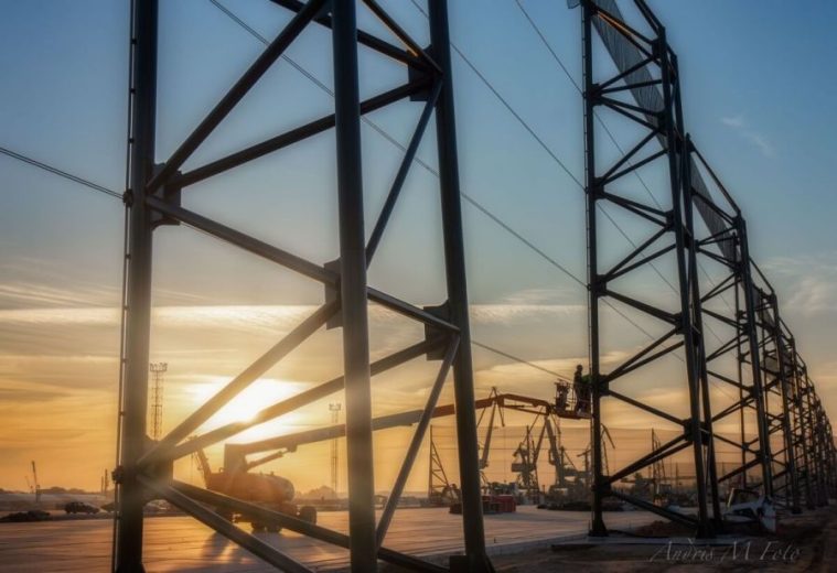 A large metal structure with workers on a lift against a sunset backdrop