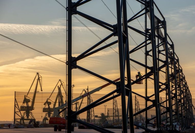 Steel framework silhouetted against a sunset with cranes and workers in the background