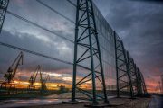 A large industrial wind fence stands against a dramatic sunset with cranes in the background