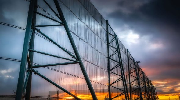 A large industrial fence structure stands against a dramatic sunset sky