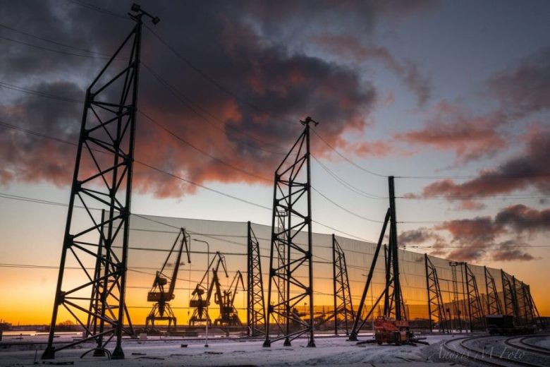 Industrial wind fences stand against a dramatic sunset with silhouetted cranes in the background