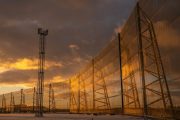 A tall metal tower stands next to a long, curved, translucent fence under a dramatic sky