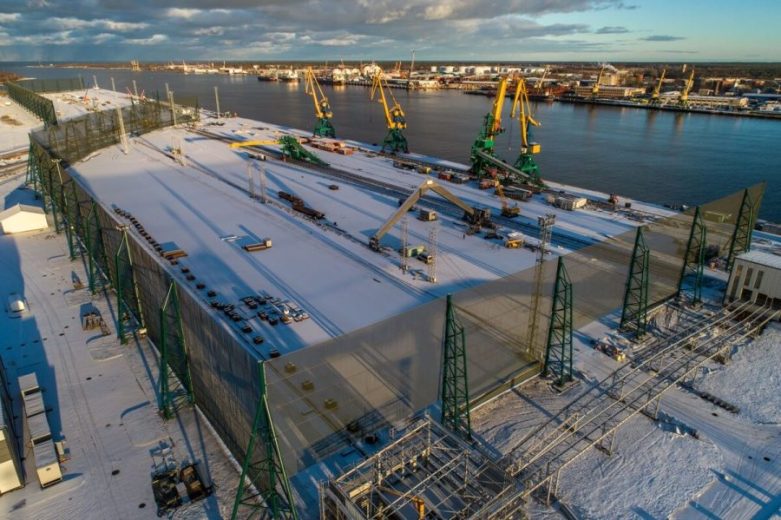A snowy industrial site with cranes and green structures along a waterfront