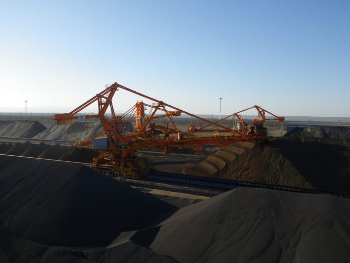Large industrial machinery operates among massive piles of dark material under a clear sky