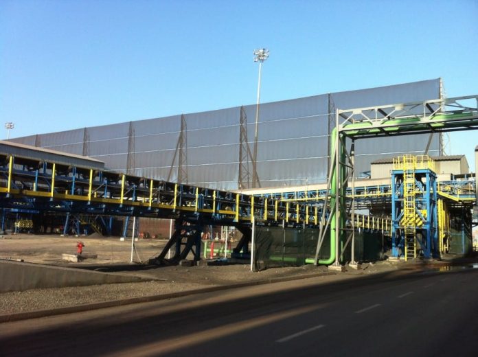 An industrial conveyor belt system with yellow and blue supports and a large building in the background