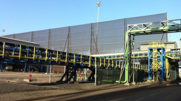 An industrial conveyor belt system with yellow and blue supports and a large building in the background