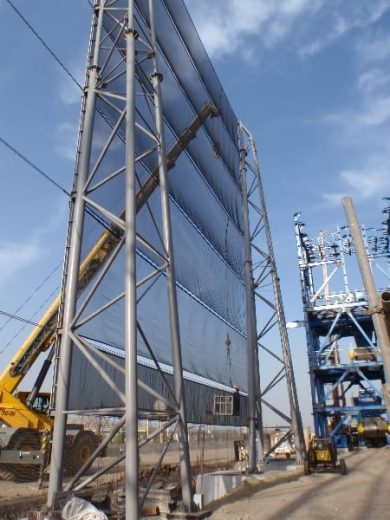 A tall metal windbreak structure stands against a clear blue sky with construction equipment nearby