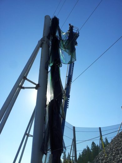 A person stands on a lift installing a green mesh netting on tall poles against a clear blue sky